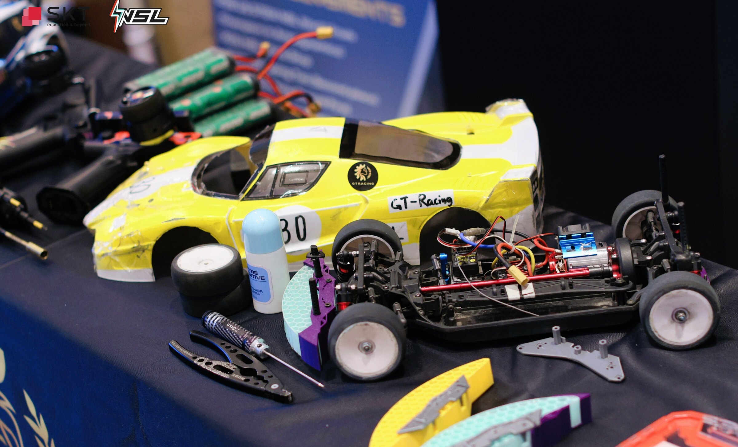 A GT Racing workbench holding the competition car, exposed chassis, tools, printed bumper parts, and tyre additive