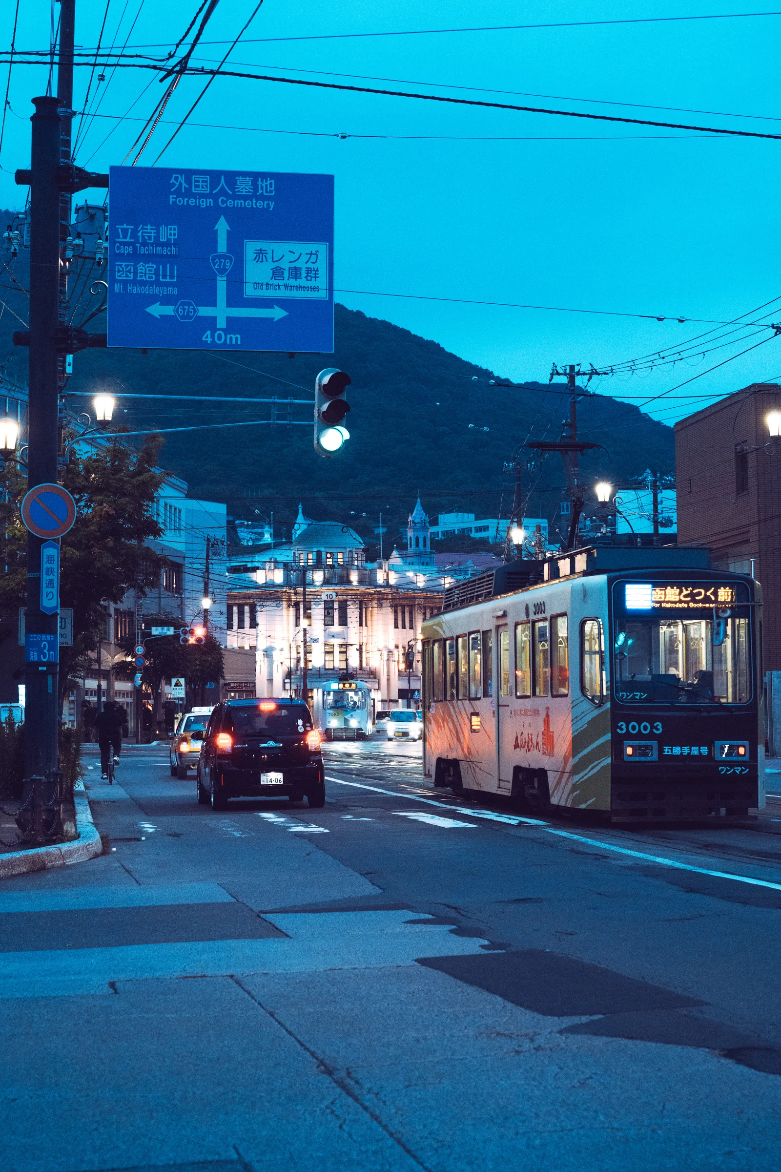 A Hakodate streetcar moving beneath overhead wires toward Mount Hakodate at blue hour