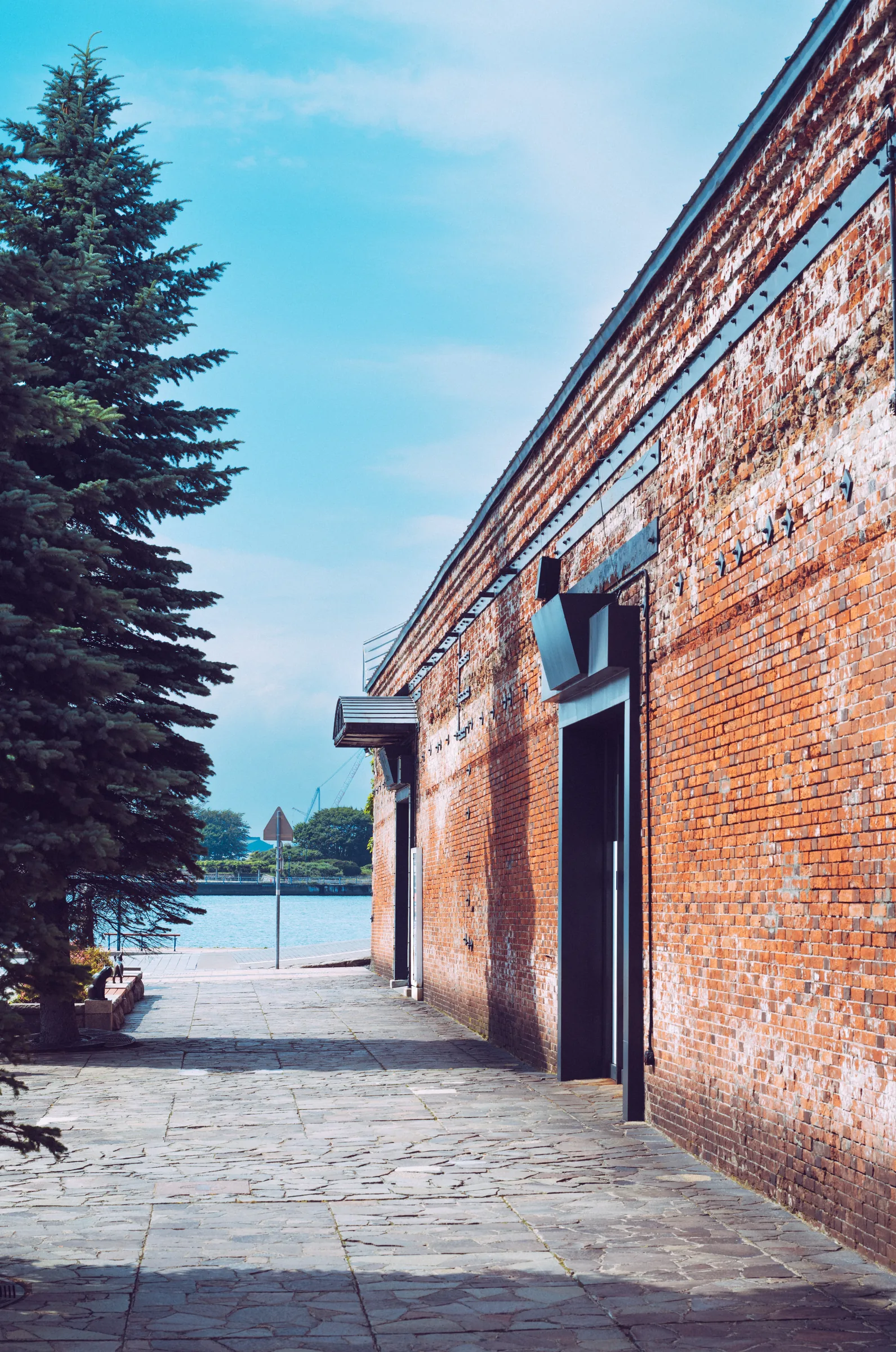 A long red-brick warehouse wall and evergreen tree opening toward Hakodate Bay