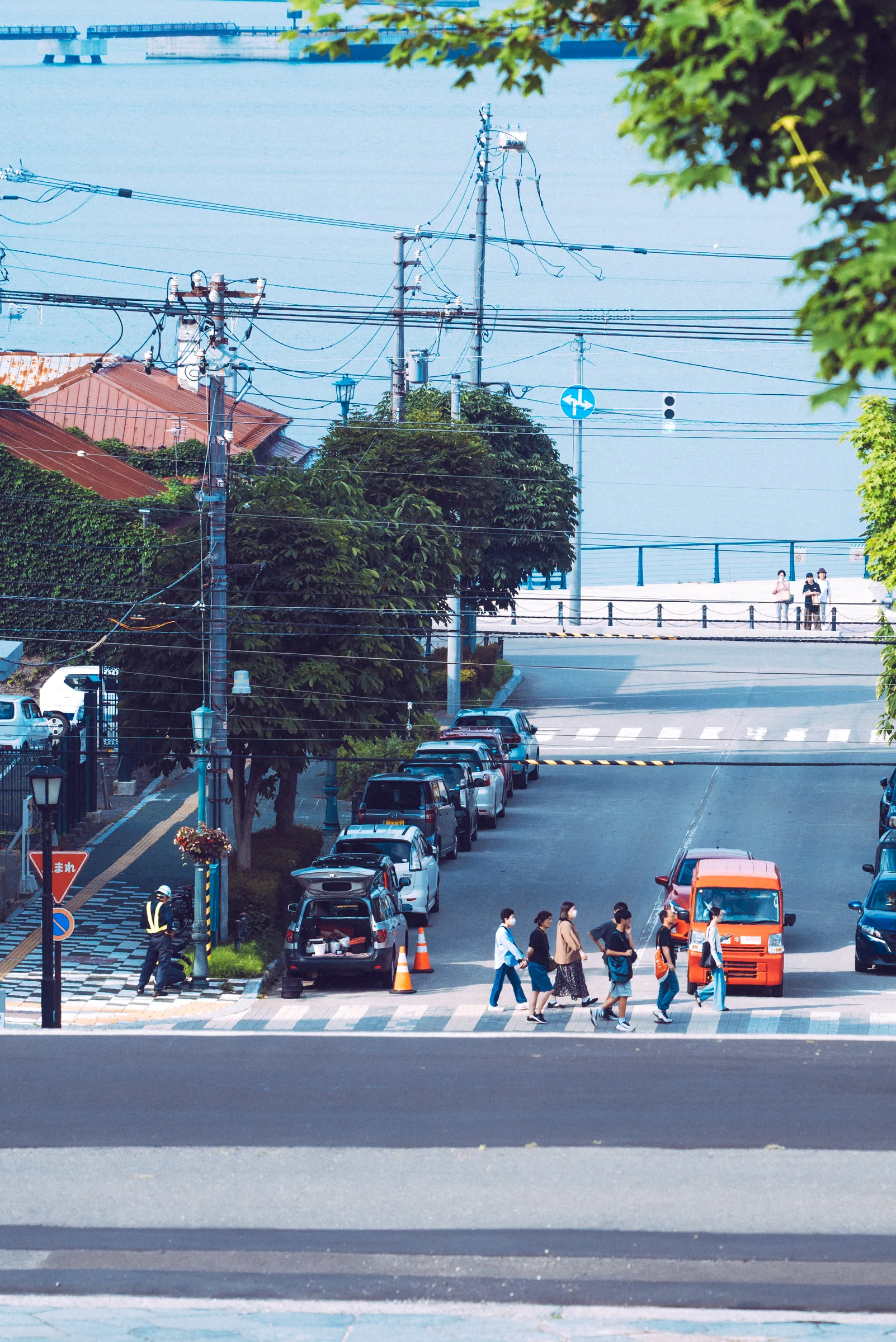 A steep Motomachi street descending through wires, pedestrians, and parked cars toward Hakodate Bay
