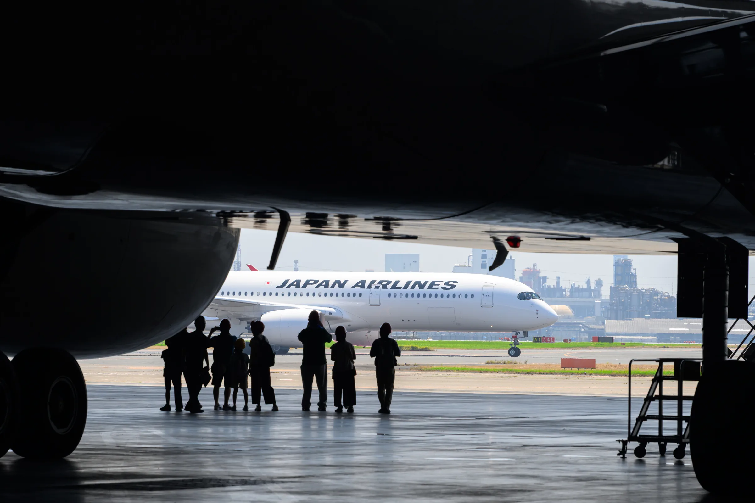 Visitors silhouetted beneath an aircraft while a Japan Airlines plane passes outside the hangar