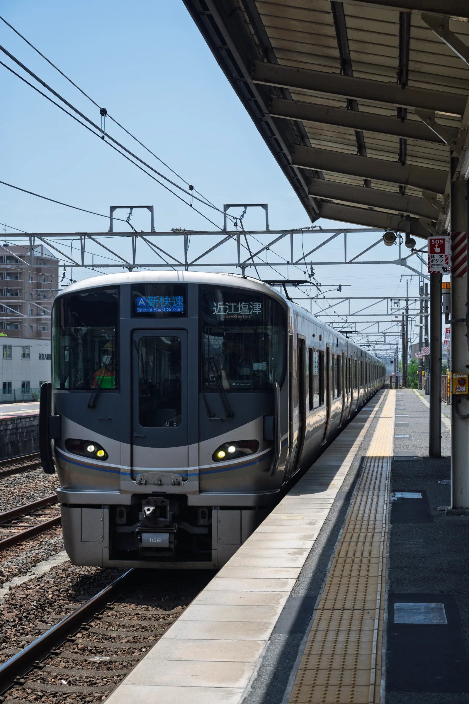A silver JR commuter train arriving beside a bright rural platform