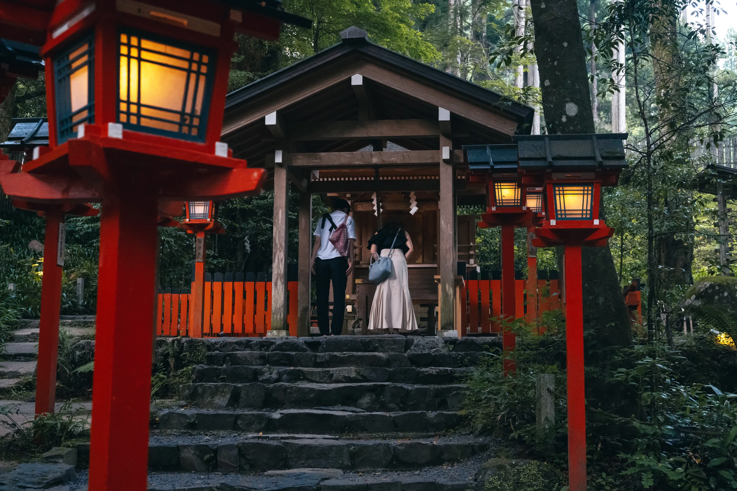 Two visitors praying at a small Kifune shrine framed by red lanterns