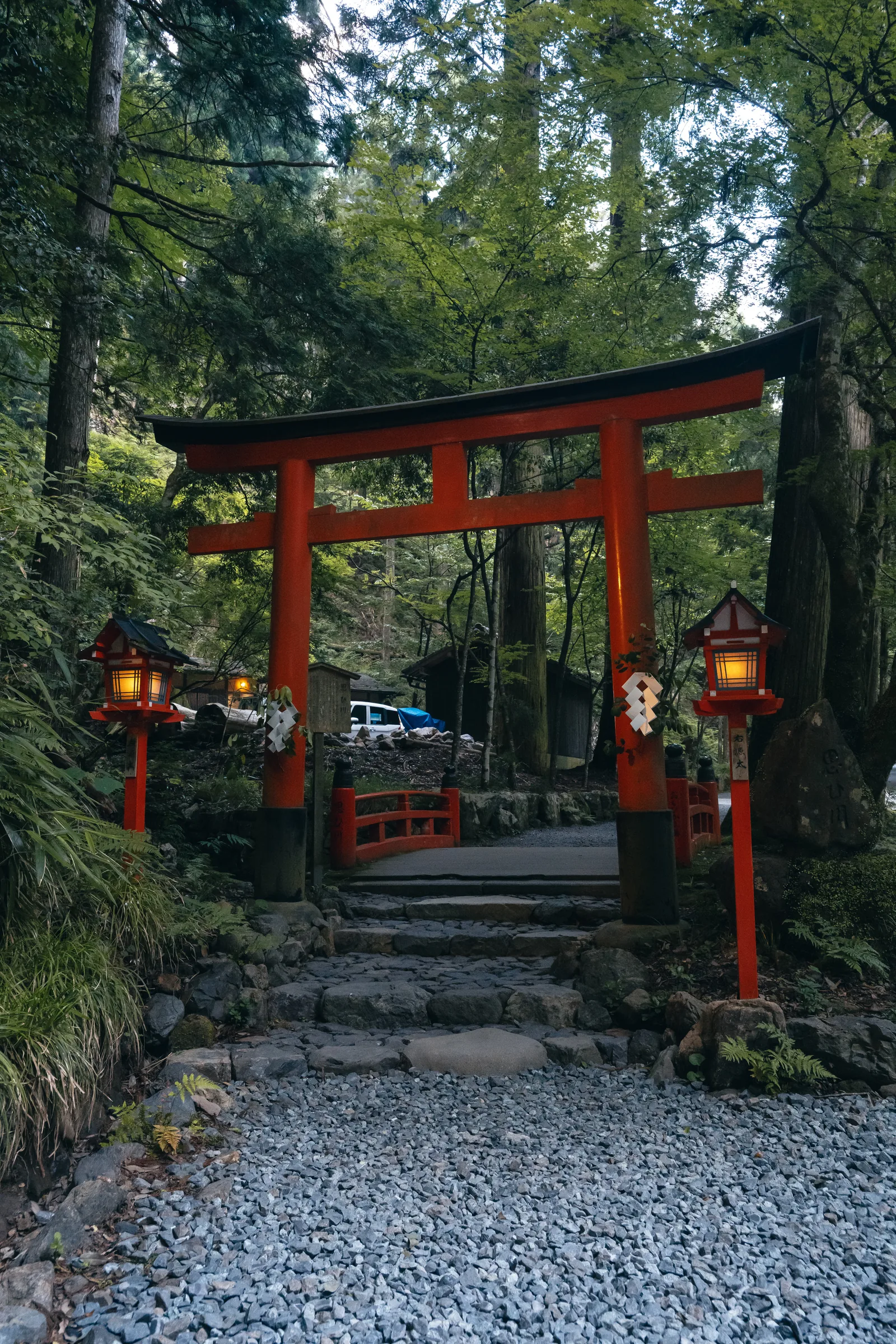 A vermilion torii and paired lanterns standing at the end of a gravel path in dense forest