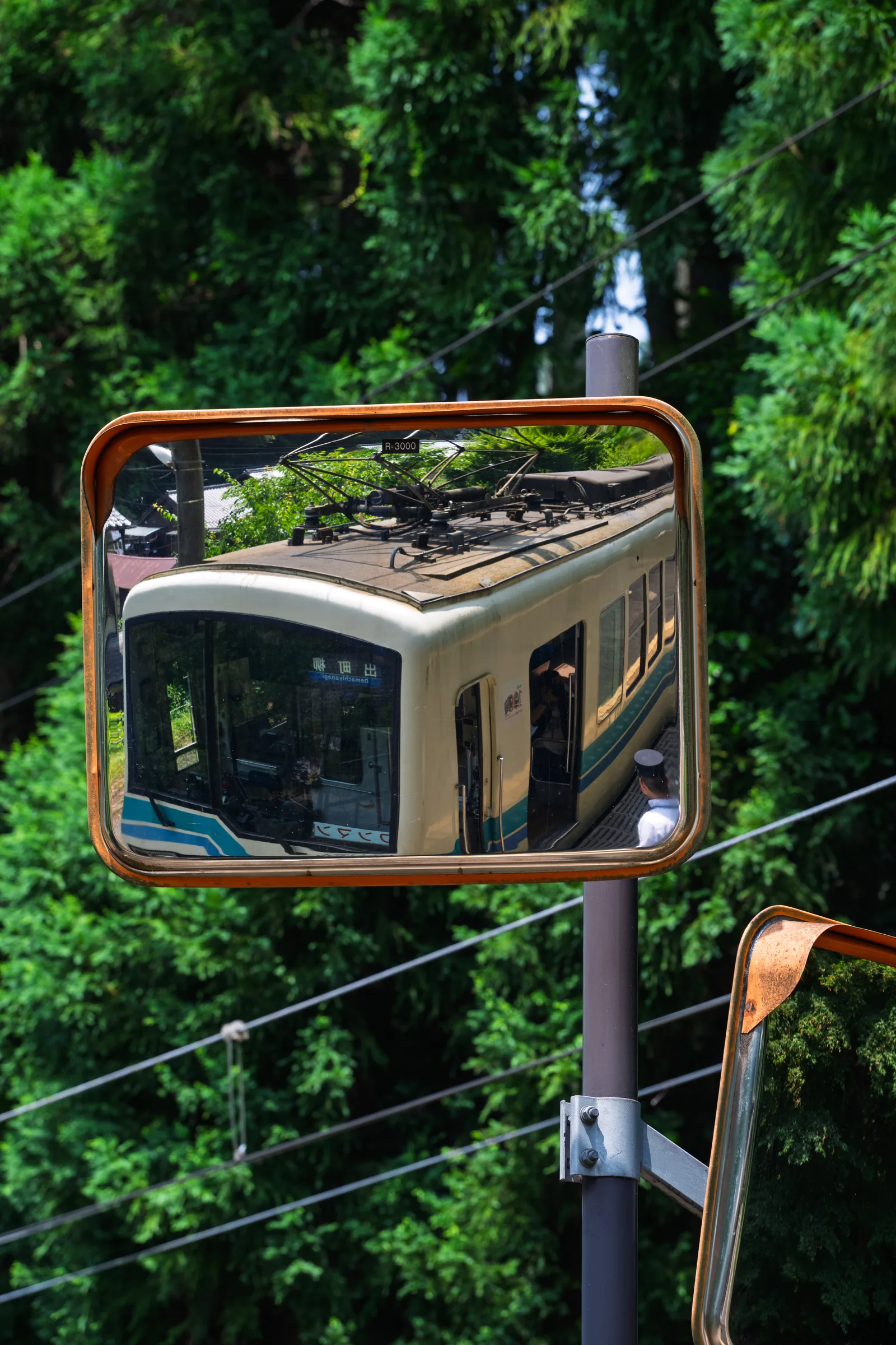 An Eizan train reflected in a convex platform mirror amid green trees