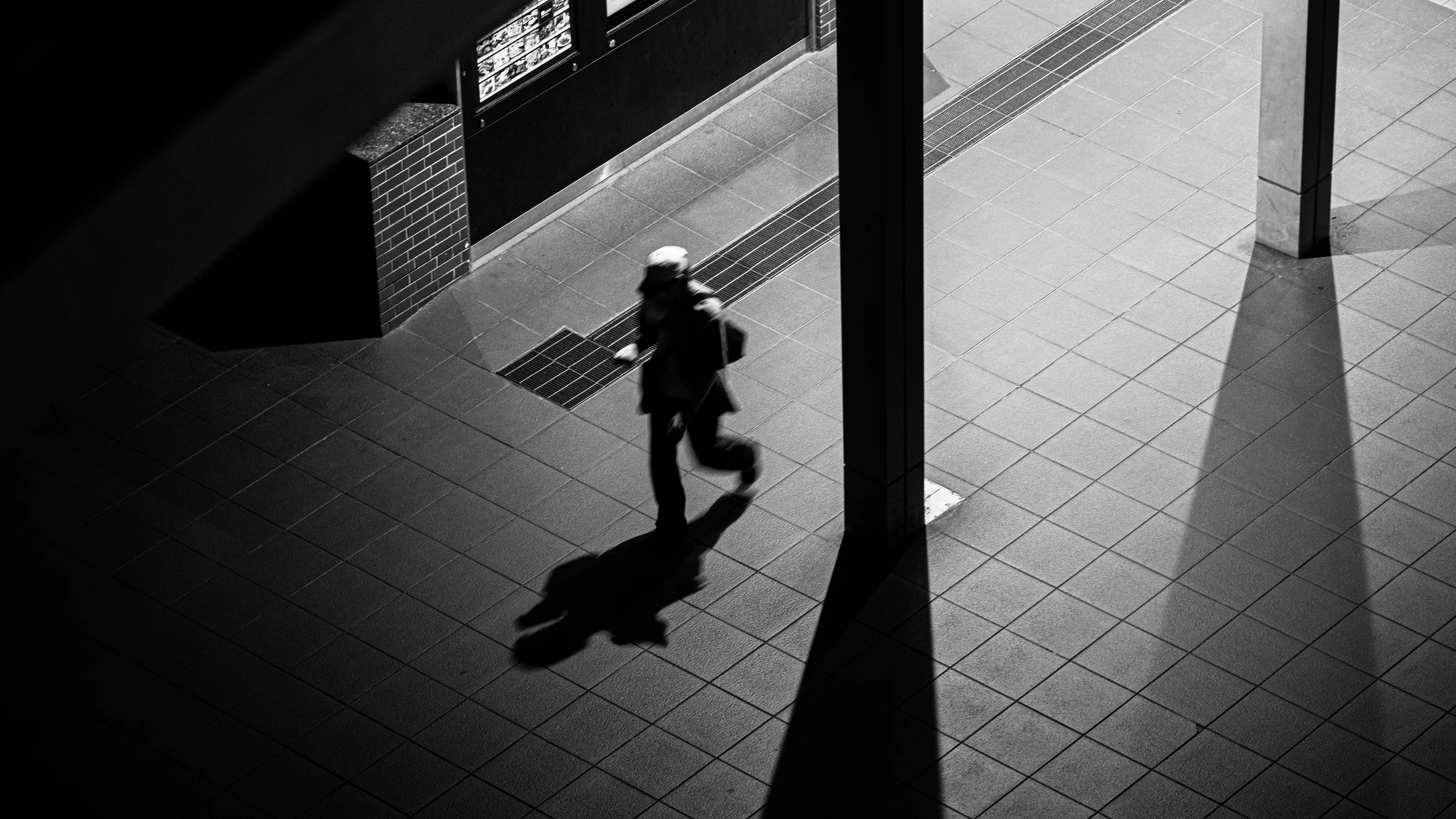 A lone traveler crossing a sharply lit station floor in black and white
