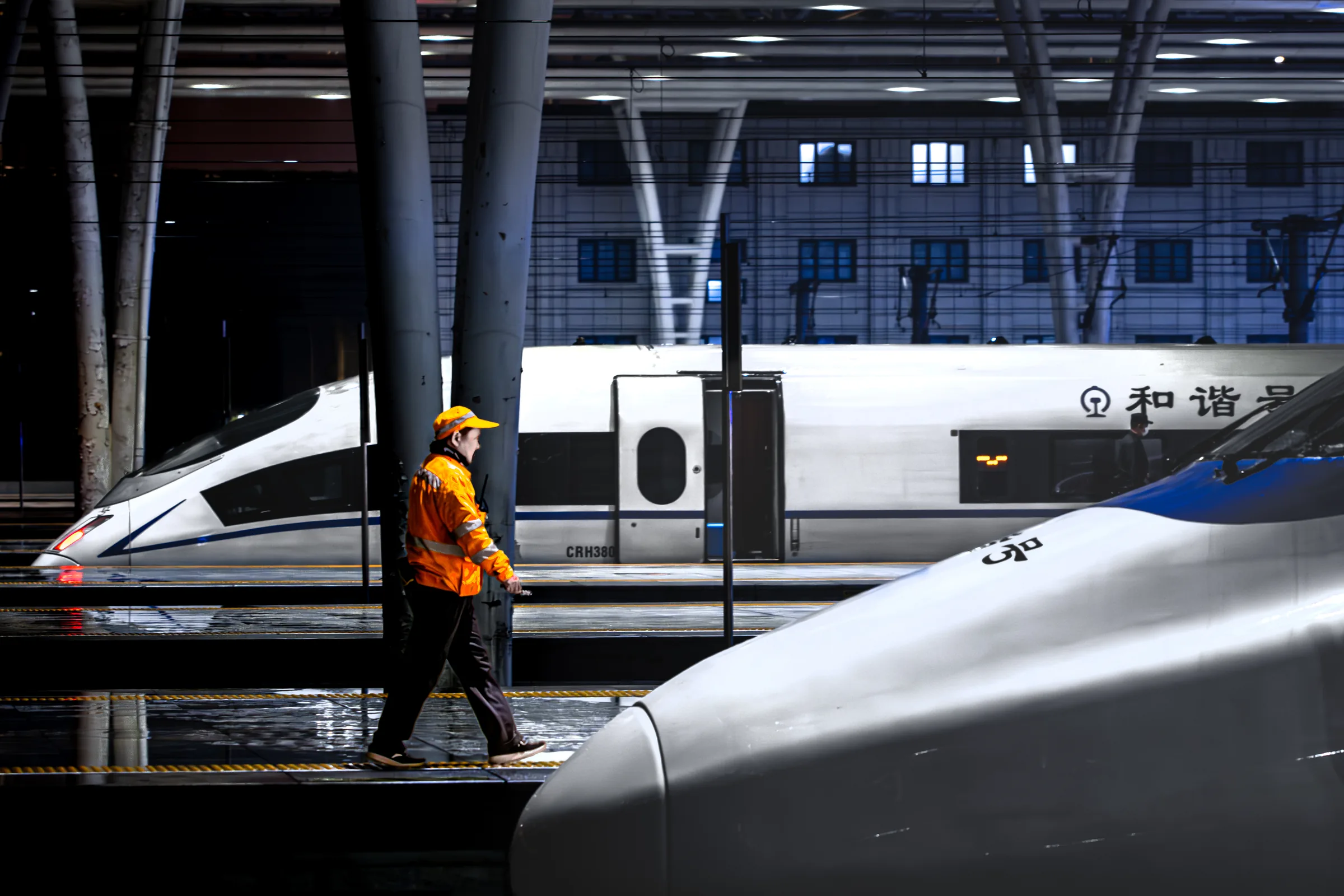 A rail worker in a bright orange jacket walking between three high-speed trains inside a station