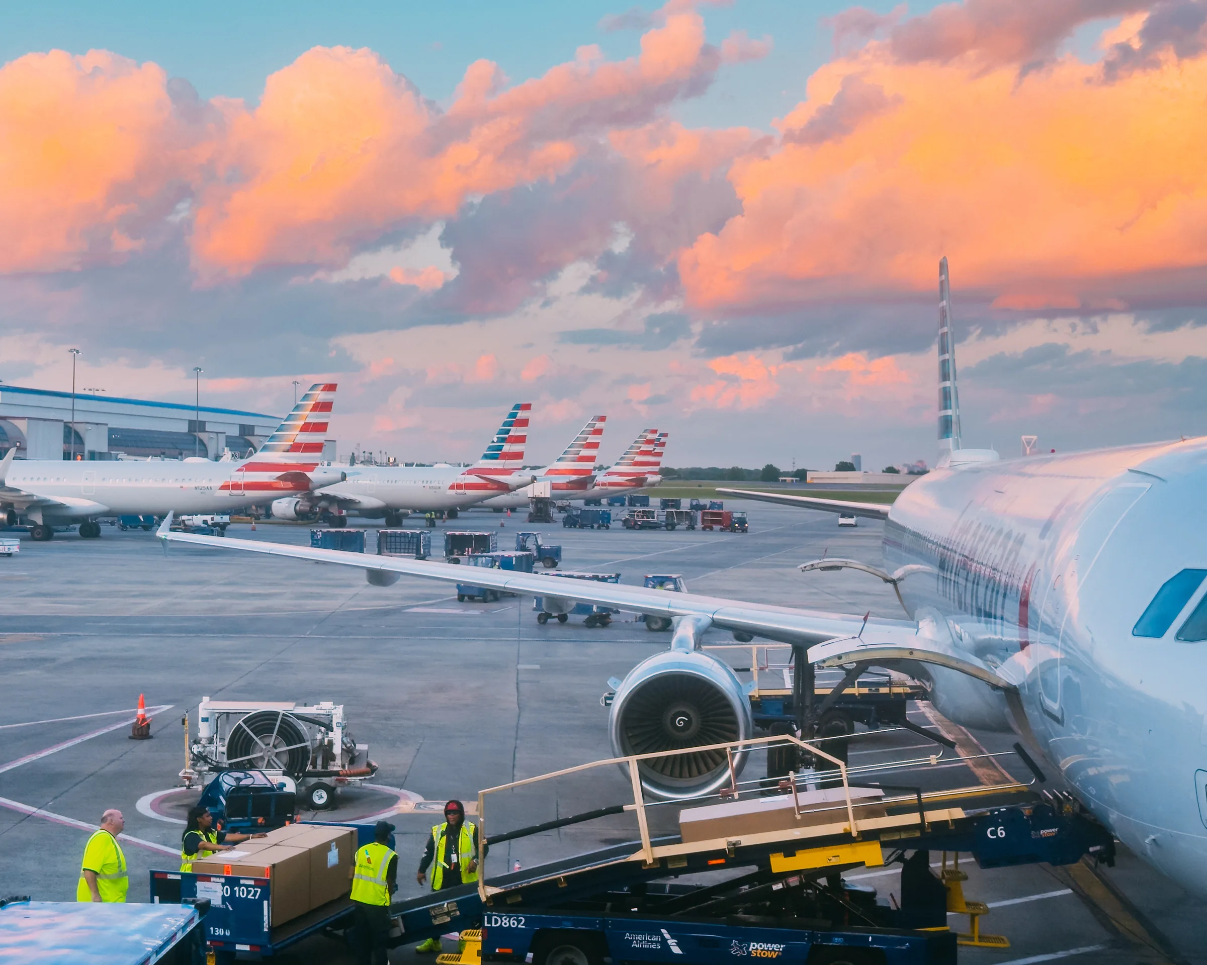 An airliner wing, engine, ground crews, and a row of American Airlines tails beneath pink storm clouds